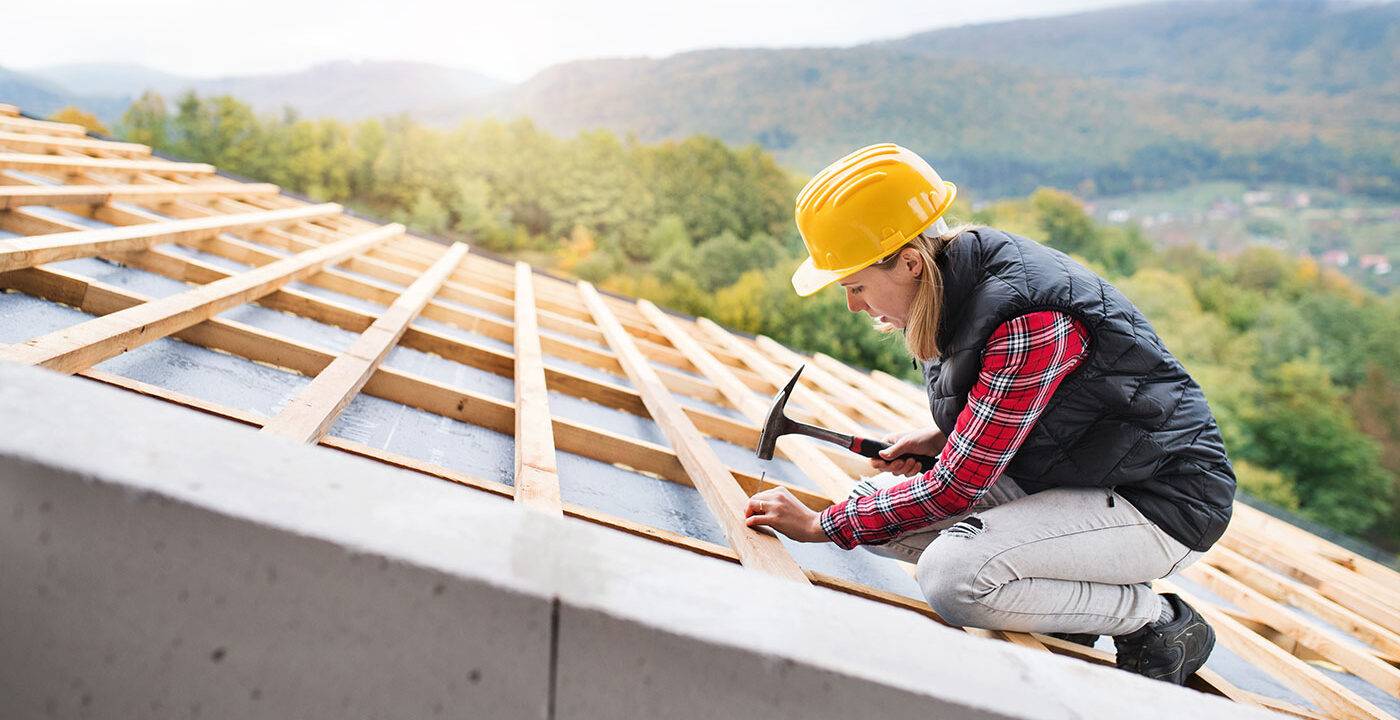 Woman roofer nailing into 2x4 on roof with a scenic mountain landscape